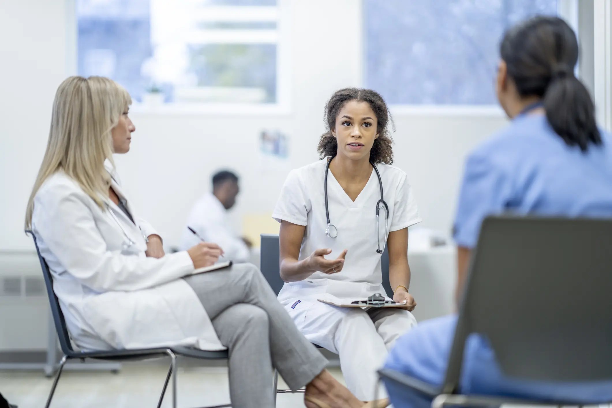 Two female medical students sit with their teacher as they go through scenarios and test their knowledge. They are wearing medical scrubs and the teacher is wearing a lab coat and has a clipboard in hand ass he takes notes.