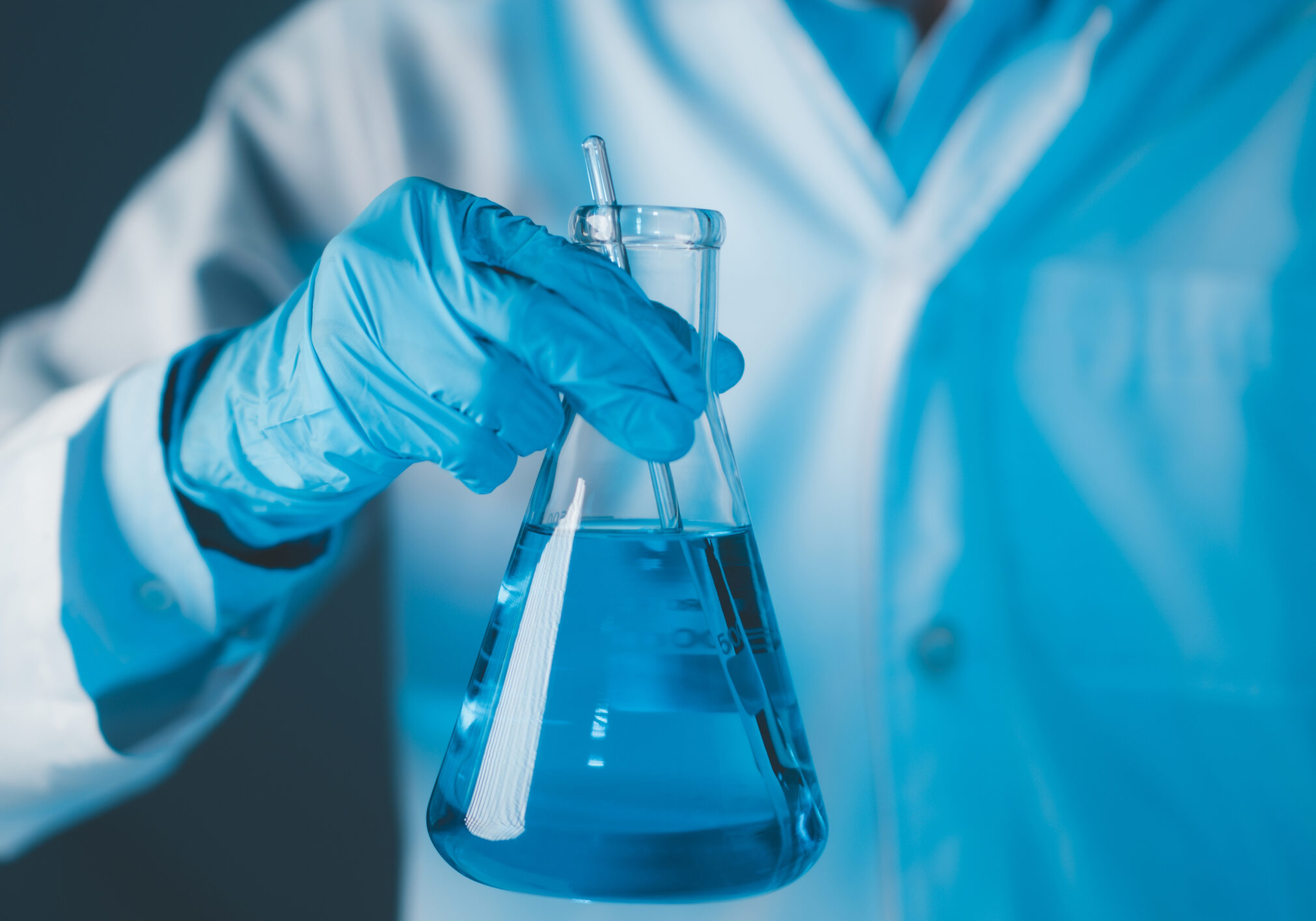 Scientist hand hold erlenmeyer flask with stirring rod filled with blue sample chemicals in chemistry science laboratory. Glassware in medical research. Scientific lab for Biotechnology.